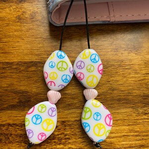 May include: A white bookmark with a peace sign pattern in yellow, pink, blue, and green. The bookmark has two pink beads and black string.