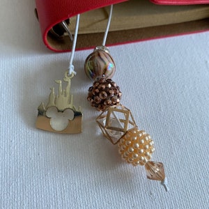 May include: A gold charm with a silhouette of a castle and Mickey Mouse ears, attached to a white string with a variety of beads, including a brown and white swirl bead, a sparkly brown bead, a clear geometric bead, and a cream-colored bead.