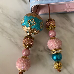 May include: Two turquoise and pink beaded bookmarks with gold accents. The turquoise bead has a floral design and the pink beads have a sparkly finish.