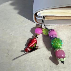 Könnte beinhalten: Ein dekorativer Lesezeichen mit einem roten Schmetterlingsanhänger und bunten Perlen. Der Schmetterling hat rot-weiße Flügel mit grünen Details. Die Perlen sind rosa, grün und lila. Das Lesezeichen ist an einem hellblauen Notizbuch befestigt.