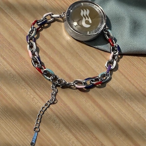 Puede incluir: Una pulsera de cadena plateada con eslabones esmaltados de colores en rojo, azul, rosa y blanco. La pulsera presenta un dije redondo plateado con texto y un logotipo. La pulsera está sobre una superficie de madera.