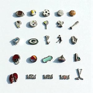 May include: A collection of silver-toned sports-themed charms. The charms depict various sports equipment and symbols, including a football, soccer ball, baseball, tennis racket, and a skateboard. The charms are arranged on a white surface.