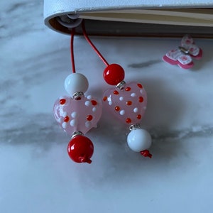 May include: Two pink heart-shaped glass beads with white and red polka dots, strung on red and white cords with white beads. The beads are hanging from a white notebook.