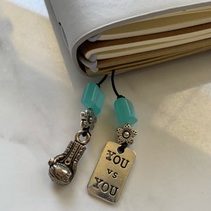 May include: A light gray notebook with a black cord bookmark featuring a boxing glove charm and a tag that reads "YOU vs YOU". The bookmark also has turquoise beads and flower accents.