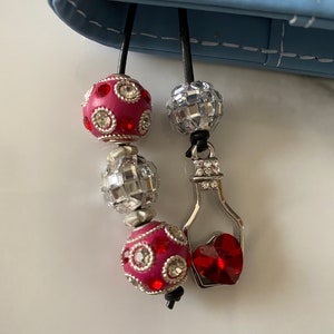May include: A beaded charm with a light blue background. The charm features two pink and silver beads with rhinestones, a disco ball bead, and a silver bottle charm with a red heart-shaped crystal. The charm is attached to a black cord.