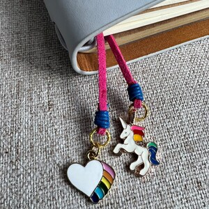 Könnte beinhalten: Zwei bunte Lesezeichen mit einem rosa und blauen Band. Ein Lesezeichen zeigt ein weißes Herz mit einem Regenbogen und das andere ein weißes Einhorn mit einer Regenbogenmähne und einem Regenbogen-Schwanz.