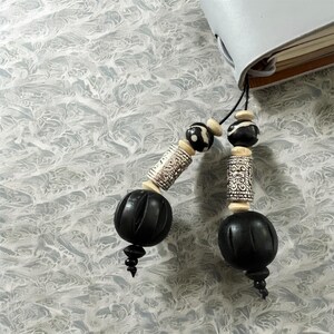 May include: Black and white beaded journal charm with a silver-colored metal accent. The charm features a large, carved black bead, a cylindrical silver bead, and smaller black and white beads. The charm is attached to a gray journal.