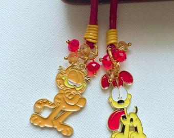 Könnte beinhalten: Zwei rote und goldene Lesezeichen-Quasten mit Comic-Figuren-Anhängern. Die Anhänger sind Garfield und Odie aus dem Garfield-Comic. Die Anhänger sind an roten Schnüren mit goldenen Perlen und roten Kristallperlen befestigt.