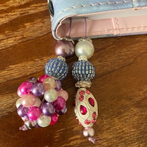 May include: A close-up of a book with a purple and blue patterned cover. A beaded bookmark with pink, purple, white, and blue beads hangs from the book.