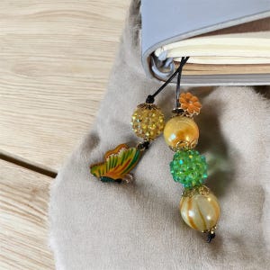 May include: A beaded bookmark with a butterfly charm. The bookmark features a series of decorative beads in yellow, green, and orange, with gold accents. A small, colorful butterfly charm is attached to the bookmark.