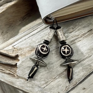 May include: Two handmade beaded bookmarks. Each bookmark features a black and white design with a cross symbol, silver-colored metal accents, and a decorative cylindrical bead. The bookmarks are attached to a book.