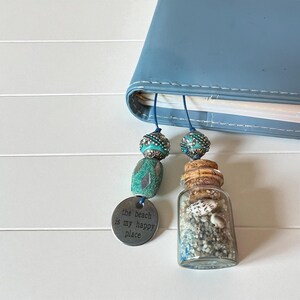 May include: A blue bookmark with turquoise beads and a small glass bottle filled with sand and seashells. A metal charm reads "the beach is my happy place." The bookmark is inserted into a blue book.
