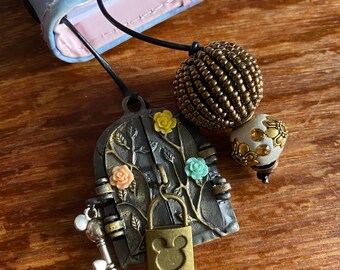 Könnte beinhalten: Ein goldfarbener Metall-Lesezeichen mit einem detaillierten Design einer Tür mit Ranken und Blumen. Die Tür hat ein Schloss in Form einer Mickey Mouse und einen Schlüssel. Das Lesezeichen enthält auch zwei dekorative Perlen, eine goldene und eine weiße mit goldenen Akzenten.