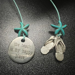 Könnte beinhalten: Eine silberne Kettenkette mit einem türkisfarbenen Seestern-Anhänger und einem silbernen Anhänger mit der Aufschrift "the beach is my happy place". Die Kette hat außerdem zwei silberne Flip-Flop-Anhänger.