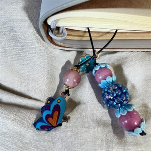 Könnte beinhalten: Zwei Perlen-Lesezeichen mit Schmetterlings- und Blumendesigns. Ein Lesezeichen hat einen blauen Schmetterlingsanhänger mit orangefarbenen und violetten Akzenten. Das andere hat rosa und blaue Perlen mit einer funkelnden blauen Perle.