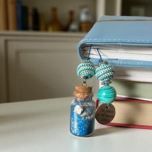 May include: A small glass bottle filled with blue sand and seashells, topped with a cork, and a beaded bookmark. The bookmark has turquoise and silver beads, and a green and white striped glass bead. A tag reads "the beach is my happy place."