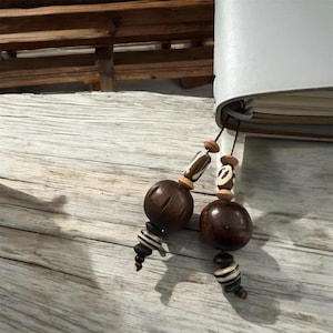 May include: A handmade bookmark with dark brown wooden beads and decorative accents. The beads are strung on a thin cord and attached to a gray leather-bound journal. The wooden beads have a natural, organic look.