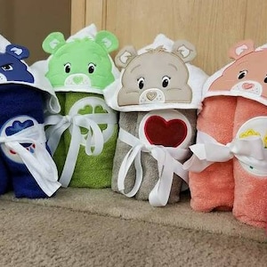 May include: A collection of hooded bath towels in various colors, each featuring a different Care Bear character design. The towels are yellow, blue, green, tan, peach, and pink, with embroidered details and tied with white ribbons.