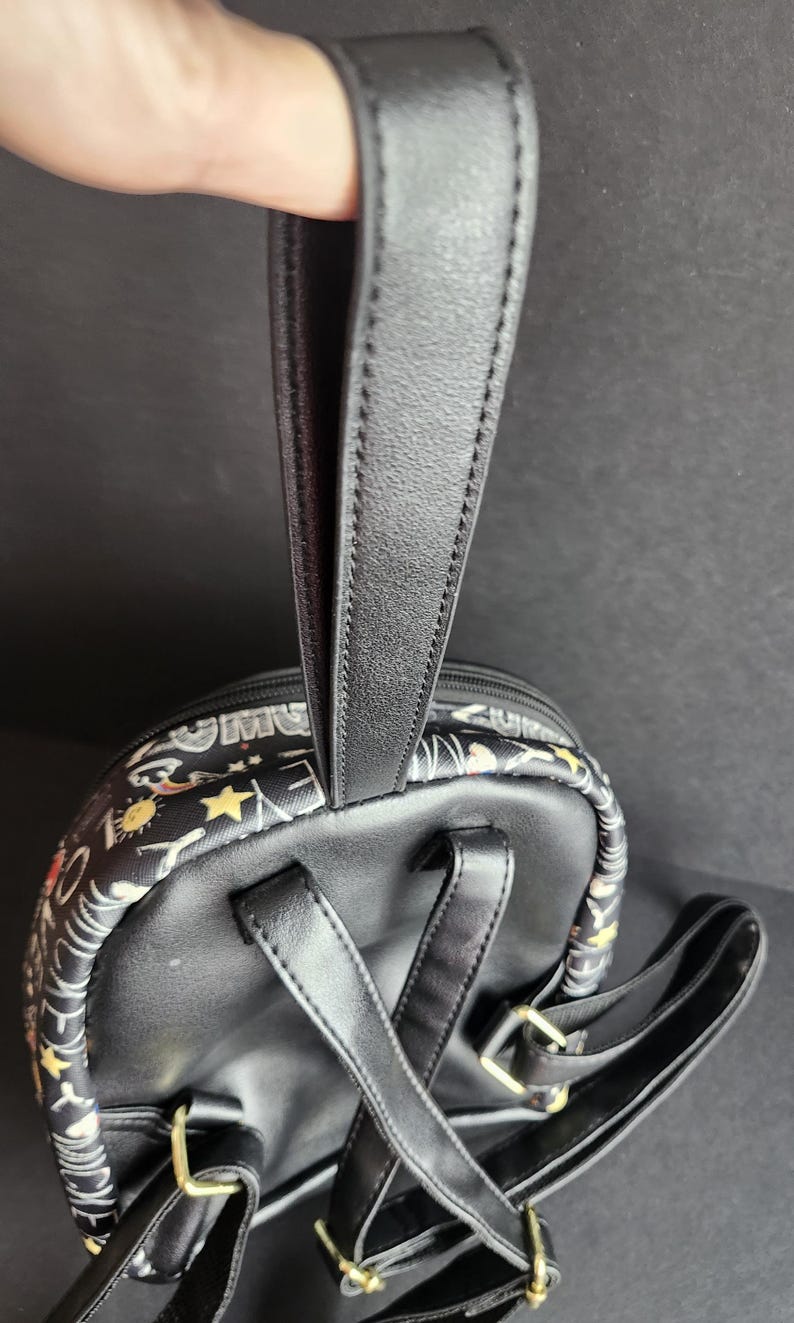 Peut inclure: Mini sac &agrave; dos noir avec une poign&eacute;e sup&eacute;rieure et des bretelles r&eacute;glables. Le sac &agrave; dos pr&eacute;sente un motif d'&eacute;toiles, de soleils et d'autres motifs. Les bretelles et la poign&eacute;e sont noires.