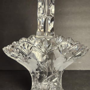 Op de afbeelding: Een heldere, geslepen glazen mand met een handvat. De mand heeft geëtste bloemmotieven en een geschulpte rand. De handgreep is rechthoekig met een gesneden patroon. De mand wordt waarschijnlijk voor decoratieve doeleinden gebruikt.