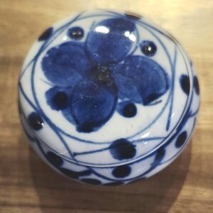 May include: A small, round, ceramic trinket box with a lid. The box is white with a blue floral design on the top and sides. The floral design includes a large flower in the center and smaller dots around the edges. The box is likely used for storing small items.