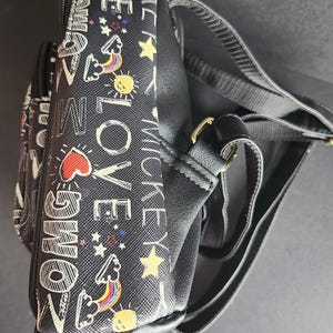 Peut inclure: Sac bandouli&egrave;re noir avec un motif r&eacute;p&eacute;t&eacute; de texte blanc, de c&oelig;urs, d'&eacute;toiles, d'arcs-en-ciel et de soleils. Le sac a une sangle noire et une quincaillerie dor&eacute;e. Le texte comprend les mots "OMG", "LOVE" et "MICKEY".