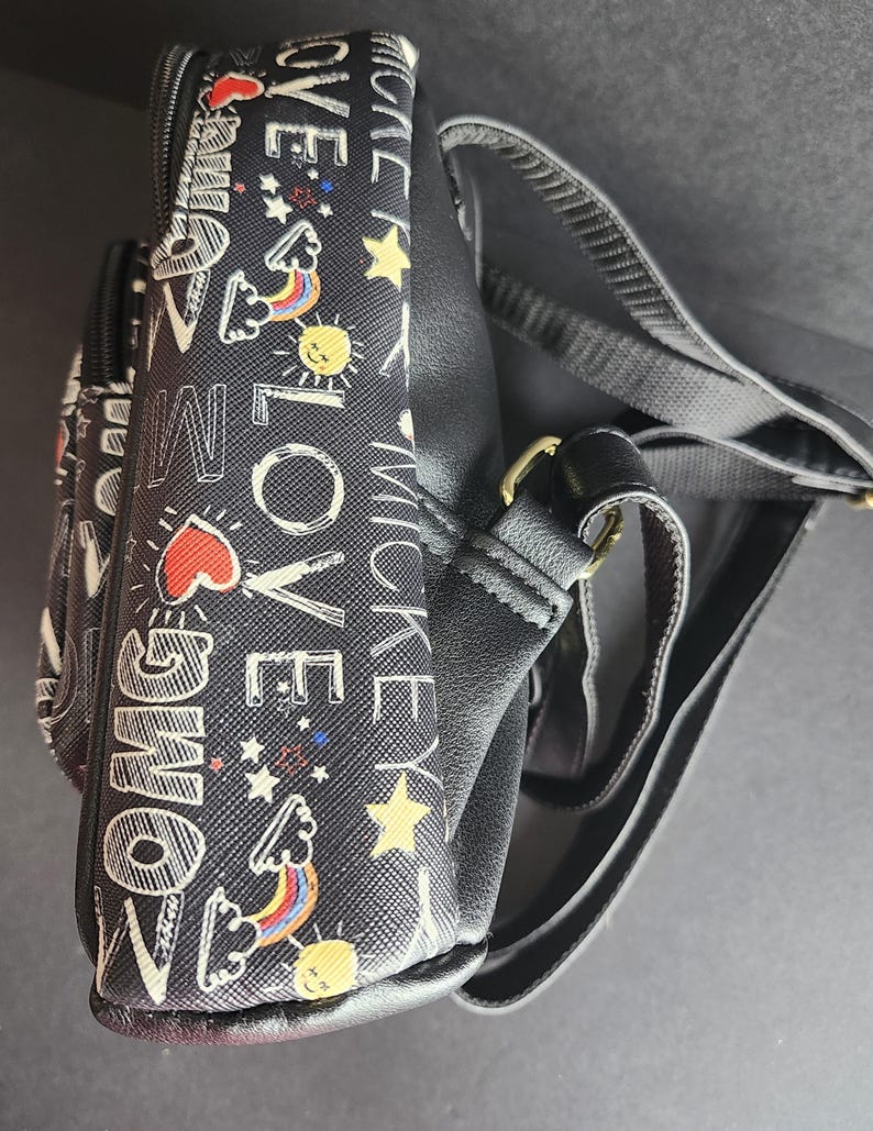 Peut inclure: Sac bandouli&egrave;re noir avec un motif r&eacute;p&eacute;t&eacute; de texte blanc, de c&oelig;urs, d'&eacute;toiles, d'arcs-en-ciel et de soleils. Le sac a une sangle noire et une quincaillerie dor&eacute;e. Le texte comprend les mots "OMG", "LOVE" et "MICKEY".