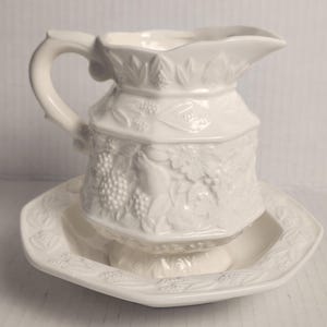 May include: A white ceramic pitcher and matching dish set. The pitcher has a curved handle and a spout. Both pieces are decorated with embossed grape and leaf designs. The dish is octagonal.
