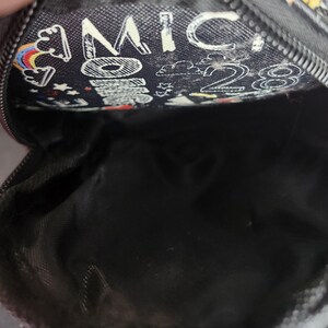 Peut inclure: Petite pochette noire zipp&eacute;e avec un motif fantaisiste. La pochette pr&eacute;sente un motif d'arcs-en-ciel, de nuages, d'&eacute;toiles et le texte "MICKEY 28" en blanc. L'int&eacute;rieur est noir.