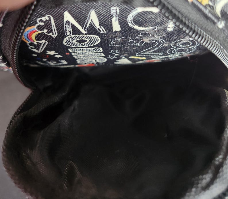 Peut inclure: Petite pochette noire zipp&eacute;e avec un motif fantaisiste. La pochette pr&eacute;sente un motif d'arcs-en-ciel, de nuages, d'&eacute;toiles et le texte "MICKEY 28" en blanc. L'int&eacute;rieur est noir.