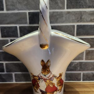 May include: White ceramic basket with a handle, featuring a Mrs. Rabbit illustration and gold trim. The basket has a flared shape and a twisted handle with gold accents. The base of the basket has the text "Mrs. Rabbit".