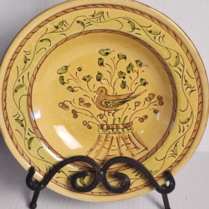 Könnte beinhalten: Dekorative Keramikplatte mit gelbem Hintergrund und braunem Rand. Die Platte zeigt einen Vogel, der auf einer Blumengesteck in der Mitte sitzt. Grüne und braune Akzente zieren den Rand der Platte.