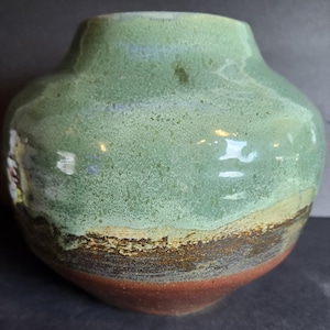 Könnte beinhalten: Eine handgefertigte Keramikvase mit rundem Körper und schmalem Hals. Die Vase hat ein geschichtetes Design mit einem grünen Oberteil, einer braunen und beigen Mitte und einem rotbraunen Boden. Die strukturierte Oberfläche trägt zu ihrem einzigartigen, künstlerischen Reiz bei.