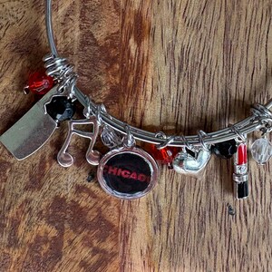 May include: Silver charm bracelet with a variety of charms. Charms include a cleaver, musical note, and a charm with "CHICAGO" in red. The bracelet also has red and black beads, a heart, and a lipstick charm.