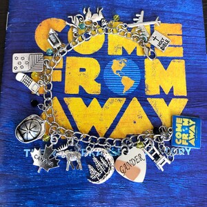May include: A silver charm bracelet with charms representing the musical "Come From Away." Charms include a plane, a heart with the word "Gander" on it, a moose, a maple leaf, a ladder, a crescent moon with trees, a fire hat, a lantern, a compass, a Holy Bible, and a square charm with the musical's title.
