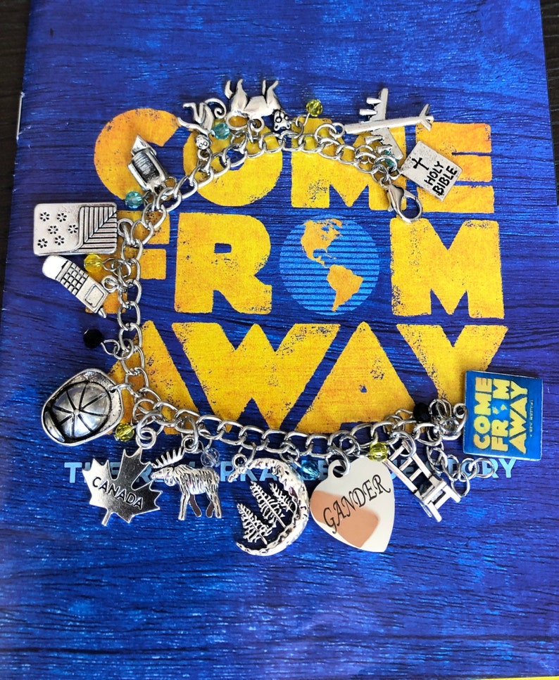 May include: A silver charm bracelet with charms representing the musical "Come From Away." Charms include a plane, a heart with the word "Gander" on it, a moose, a maple leaf, a ladder, a crescent moon with trees, a fire hat, a lantern, a compass, a Holy Bible, and a square charm with the musical's title.