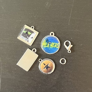May include: Six silver charms with different musical show logos. The charms are: Wicked, Heathers, Hamilton, a blank rectangle, a silver jump ring, and a silver lobster clasp.