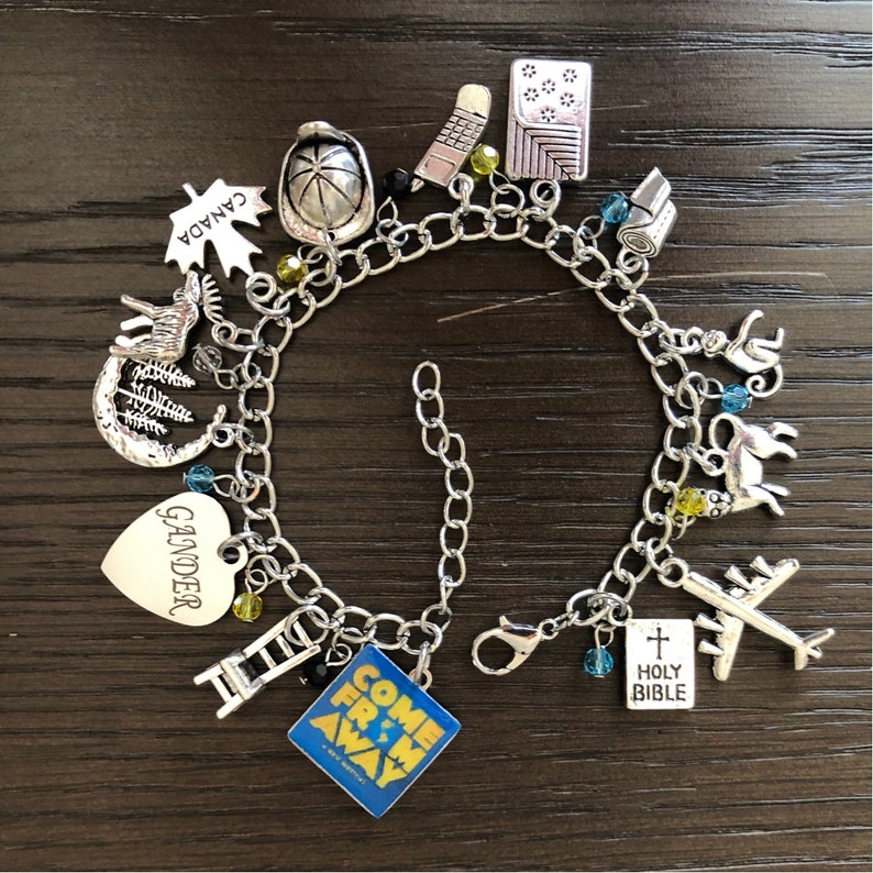 May include: A silver charm bracelet with various charms, including a heart charm with the word "Gander" on it, a charm with the words "Come From Away" on it, a charm with a maple leaf and the word "Canada" on it, a charm with a cross and the words "Holy Bible" on it, and a charm with a plane on it.