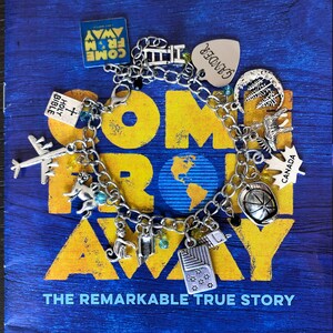 May include: A silver charm bracelet with various charms, including a plane, a dog, a book, a ladder, a heart with the word "Gander" on it, a maple leaf with the word "Canada" on it, a basketball, and a book with the words "Holy Bible" on it. The bracelet is on a book with the words "Come From Away" and "The Remarkable True Story" on it.