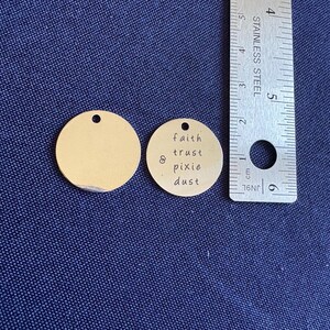 May include: Two gold-colored metal round tags. One tag is blank and the other tag has the text "faith & trust pixie dust" engraved on it.