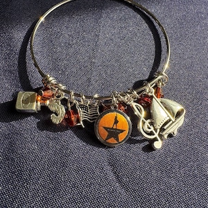 Peut inclure: Un bracelet à breloques en argent avec plusieurs breloques, dont une note de musique, un bateau, une étoile avec le mot "Hamilton" dessus et un petit drapeau américain. Les breloques sont attachées à un bracelet en argent.