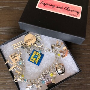 May include: A silver charm bracelet with various charms, including a "Come From Away" charm, a "Canada" charm, a "Holy Bible" charm, a ladder charm, a "Gander" charm, a maple leaf charm, a hockey puck charm, a plane charm, a musical note charm, and a building charm. The bracelet is in a black gift box with a pink and white label that says "Inspiring and Charming Etsy Shop".