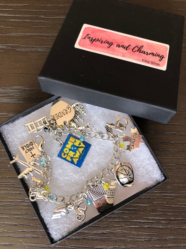 May include: A silver charm bracelet with various charms, including a "Come From Away" charm, a "Canada" charm, a "Holy Bible" charm, a ladder charm, a "Gander" charm, a maple leaf charm, a hockey puck charm, a plane charm, a musical note charm, and a building charm. The bracelet is in a black gift box with a pink and white label that says "Inspiring and Charming Etsy Shop".