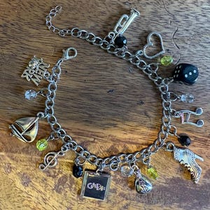 May include: Silver charm bracelet with various charms, including a sailboat, musical notes, a trumpet, a heart, a dice, and a money bag. The bracelet also features charms that say "New York" and "G&Ds". The charms are accented with black and green beads.