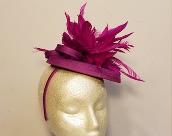 May include: A magenta fascinator hat with a headband, displayed on a mannequin head. The hat features a layered design and is adorned with magenta feathers, creating an elegant accessory for special occasions.