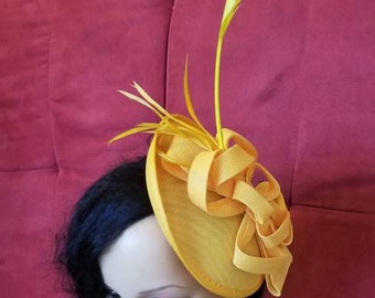 May include: A yellow fascinator with a large bow and feathers. The fascinator is worn by a person with black hair and a black shirt.