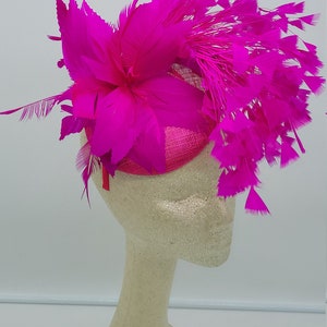 May include: A bright pink feather hat with a large flower and a small, round base. The hat is displayed on a white mannequin head.