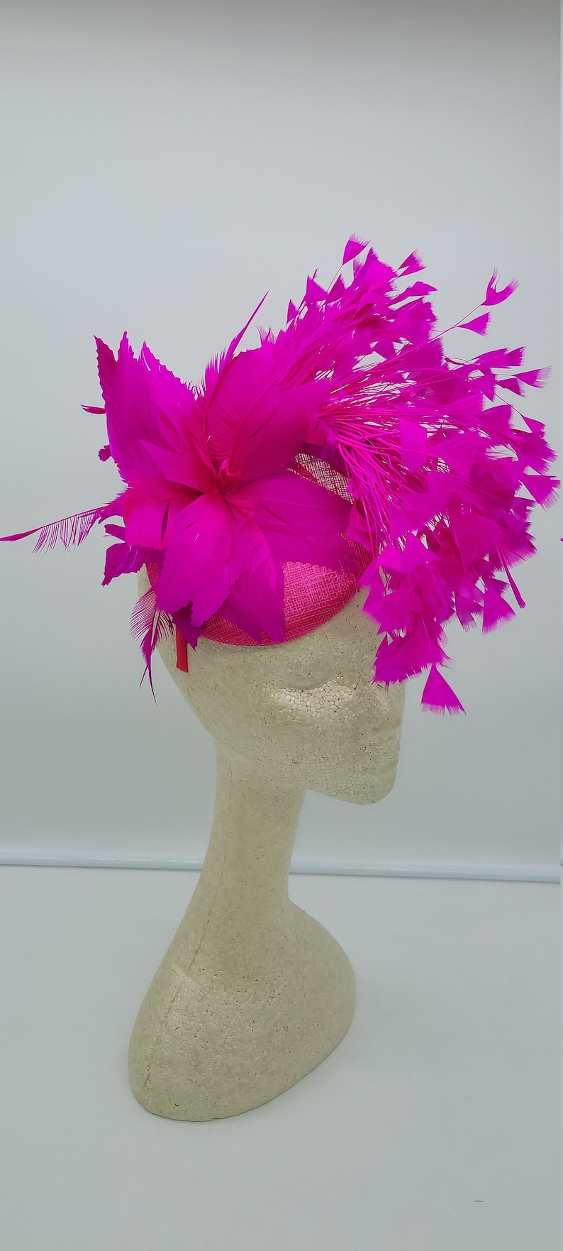 May include: A bright pink feather hat with a large flower and a small, round base. The hat is displayed on a white mannequin head.
