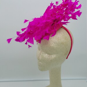 May include: A bright pink feather headband with a large, fluffy feather design. The headband is worn on a white mannequin head.