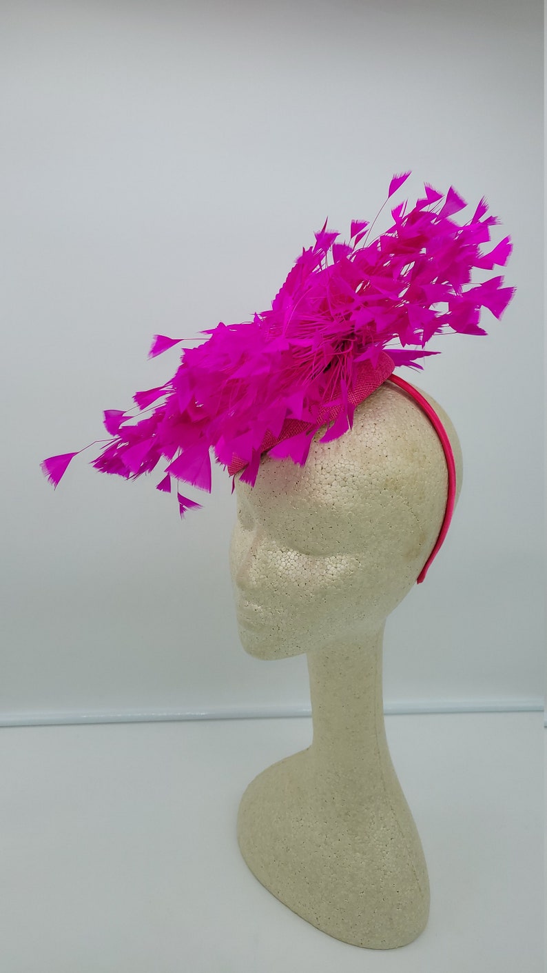 May include: A bright pink feather headband with a large, fluffy feather design. The headband is worn on a white mannequin head.